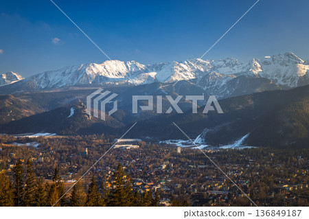 Majestic snow-capped mountains tower over a sprawling town nestled in a valley, beneath a clear blue winter sky. Zakopane town at the foot of the Tatra Mountains in Poland, Europe. 136849187