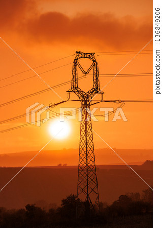 A towering electricity pylon silhouette stands majestically against a blazing orange sky at sunset. Power lines stretch across a scenic, rural landscape. 136849206