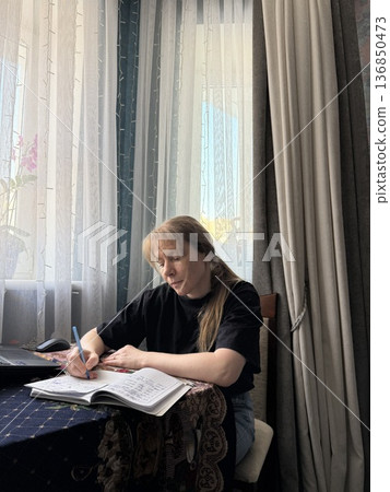 Focused woman writing in a notebook while sitting at a table by the window Focused woman writing in a notebook while sitting at a table by the window 136850473