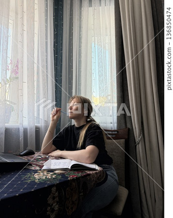 Thoughtful woman sitting at a table over her notes with a pen at her chin and looking out the window Thoughtful woman sitting at a table over her notes with a pen at her chin and looking out the window 136850474