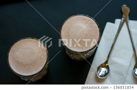 Top view of two glasses of creamy hot cocoa with smooth milk foam, served with spoons and napkin. Cozy cafe atmosphere and warm drink concept. Top view of two glasses of creamy hot cocoa with smooth milk foam, served with spoons and napkin. Cozy cafe atmosphere and warm drink concept. 136851854