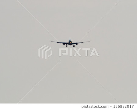 Large passenger aircraft with landing gear extended descends for landing. The modern jet is centered in the frame against a plain light grey sky, showing a daytime approach to an airport. Large passenger aircraft with landing gear extended descends for landing. The modern jet is centered in the frame against a plain light grey sky, showing a daytime approach to an airport. 136852017