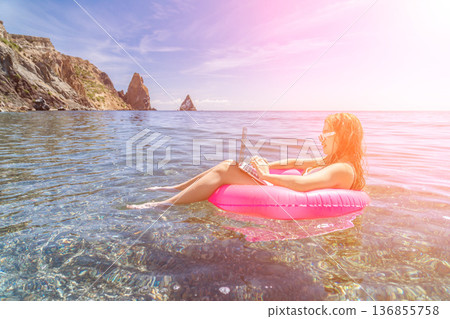 Remote work digital nomad freelance woman sitting on pink inflatable ring using laptop while floating in clear blue ocean water under sun flare Remote work digital nomad freelance woman sitting on pink inflatable ring using laptop while floating in clear blue ocean water under sun flare 136855758