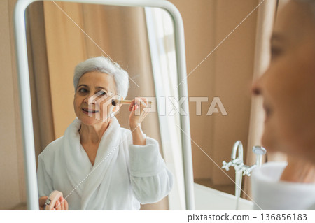 Mature adult old woman with smooth soft perfect skin applying rouge with brush on cheek looking at mirror doing daily beauty make up routine. Senior woman putting blush on her face. Anti age makeup 136856183