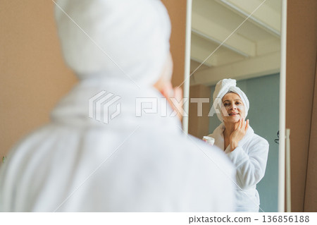 Mature adult old woman looking at mirror applying nourishing anti age face cream doing daily beauty routine. Senior woman with white mask creme on skin gets facial treatment Skin care aging skincare Mature adult old woman looking at mirror applying nourishing anti age face cream doing daily beauty routine. Senior woman with white mask creme on skin gets facial treatment Skin care aging skincare 136856188