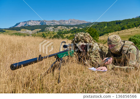 Camouflaged Military Sniper And Spotter Lying In Tall Grass With Rifle In Mountain Field 136856566