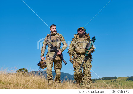 Two Soldiers in Camouflage and Tactical Gear Walking Through Open Field With Rifles 136856572