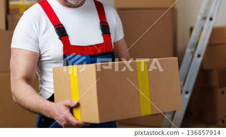Worker carries a large cardboard box in a warehouse during the day Worker carries a large cardboard box in a warehouse during the day 136857914
