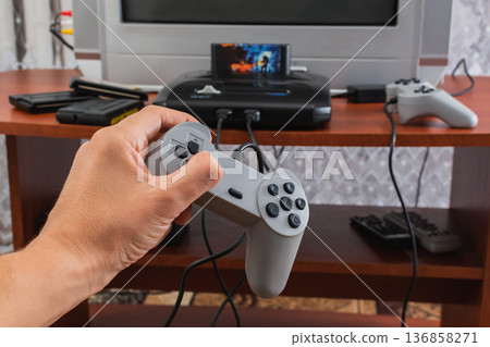 Passion for computer and console games. The process of playing games in a home environment. Hand holding a game controller Passion for computer and console games. The process of playing games in a home environment. Hand holding a game controller 136858271