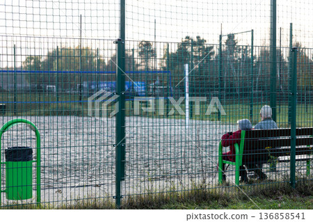 Elderly couple sitting on bench watching volleyball game at local court Elderly couple sitting on bench watching volleyball game at local court 136858541