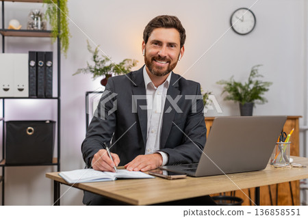 Middle-aged businessman attends webinar on laptop wears earphones takes notes snaps smartphone photo Middle-aged businessman attends webinar on laptop wears earphones takes notes snaps smartphone photo 136858551