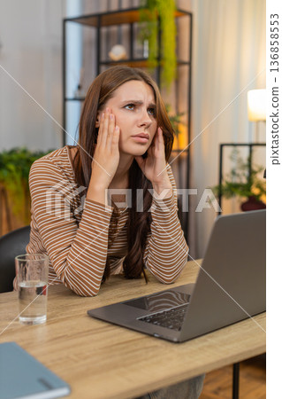 Young woman rubs temples after long laptop work drinks water squints from fatigue and breathes slow Young woman rubs temples after long laptop work drinks water squints from fatigue and breathes slow 136858553