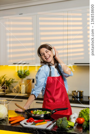 Young woman dancing to music in headphones cooking healthy vegetables in home kitchen for diet meal Young woman dancing to music in headphones cooking healthy vegetables in home kitchen for diet meal 136858600