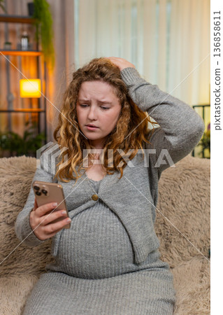 Pregnant young woman reading stressful news on smartphone, concerned expression at home on sofa Pregnant young woman reading stressful news on smartphone, concerned expression at home on sofa 136858611
