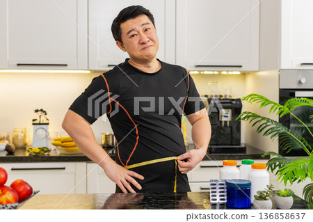 Heavy Asian adult man measures waist with tape upset about big belly fat weight gain at home kitchen Heavy Asian adult man measures waist with tape upset about big belly fat weight gain at home kitchen 136858637