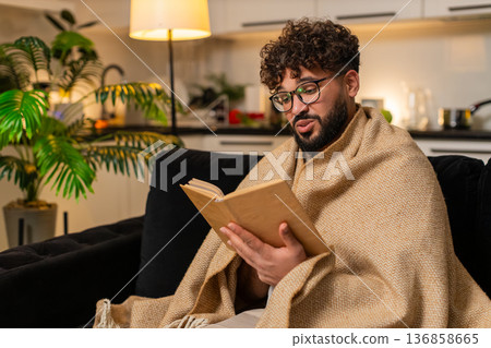 Arabian man in glasses wrapped in blanket reads novel on quiet evening relaxed mood focus calm peace Arabian man in glasses wrapped in blanket reads novel on quiet evening relaxed mood focus calm peace 136858665