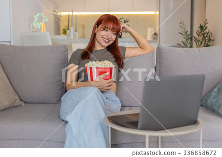 Young woman relaxes with popcorn watches funny trailer on laptop laughs smiles enjoys comedy preview Young woman relaxes with popcorn watches funny trailer on laptop laughs smiles enjoys comedy preview 136858672