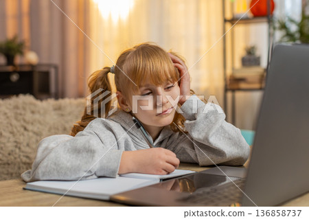 Tired young girl doing homework at table with laptop writing in notebook and showing lazy expression Tired young girl doing homework at table with laptop writing in notebook and showing lazy expression 136858737