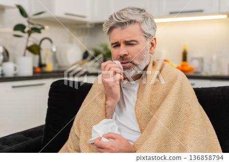 Middle-aged man wrapped in plaid coughs wipes nose with tissue feels weak flu symptoms rest 136858794