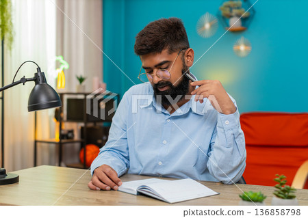 Indian man holds pen gazes notebook deeply thinking planning ideas writing strategy indoors focused 136858798