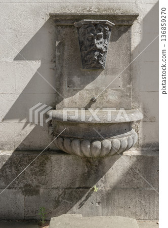 Mounted against a weathered wall at Chiswick House, a masterfully carved mascaron a bearded stone visage presides over a stone basin. Mounted against a weathered wall at Chiswick House, a masterfully carved mascaron a bearded stone visage presides over a stone basin. 136859270