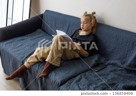 Young girl sitting on blue sofa and reading book at home. Child 6 years old Young girl sitting on blue sofa and reading book at home. Child 6 years old 136859409