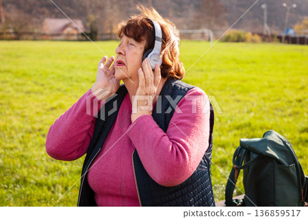 Mature Caucasian woman enjoying favorite music with headphones during relaxation in a public park. Calm nature environment supports happiness, freedom, and positive retirement experience. 136859517