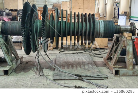 Industrial turbine rotor with multiple stages of blades rests on heavy metal supports inside a workshop, with steel cables in the foreground Industrial turbine rotor with multiple stages of blades rests on heavy metal supports inside a workshop, with steel cables in the foreground 136860293
