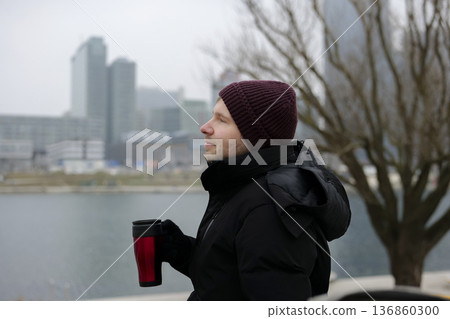 Young man enjoying a warm drink from a disposable cup near the river on a chilly day in winter. Concept of a city winter activity 136860300