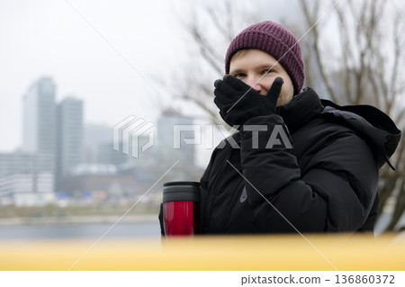 Young man enjoying a warm drink from a red travel mug near the river on a chilly day in winter. Concept of a city winter activity Young man enjoying a warm drink from a red travel mug near the river on a chilly day in winter. Concept of a city winter activity 136860372
