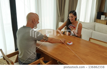 A man and a woman play cards at a wooden table in a sunlit modern living room A man and a woman play cards at a wooden table in a sunlit modern living room 136862338
