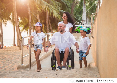 Disabled man in a wheelchair moves on a ramp to the beach with his family. 136862770