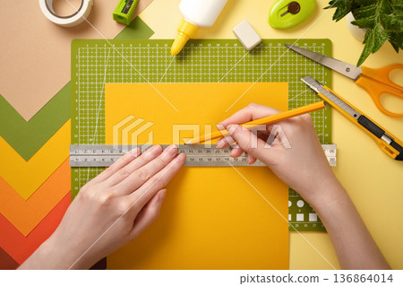 Hands draw line with pencil along ruler on yellow paper. Cutting mat, scissors, glue, and stationery. Hands draw line with pencil along ruler on yellow paper. Cutting mat, scissors, glue, and stationery. 136864014
