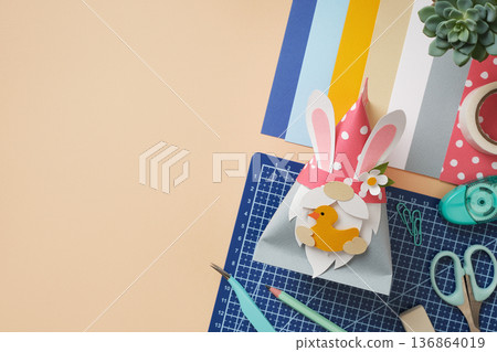 DIY Easter paper gnome with bunny ears holding a yellow duck. Creative workspace with tweezers, pencils, and colorful paper. Copyspace. 136864019