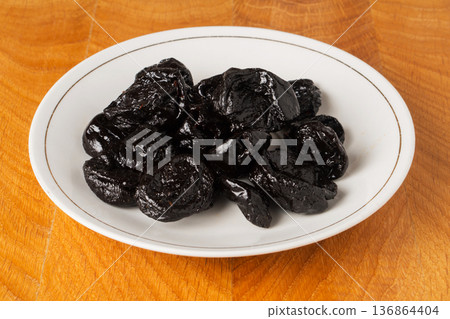 Prune dried fruit lies in a bunch in a white plate on a wooden board 136864404