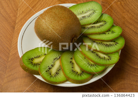 Kiwi single whole and sliced in pieces in a white plate and on a wooden board Kiwi single whole and sliced in pieces in a white plate and on a wooden board 136864423