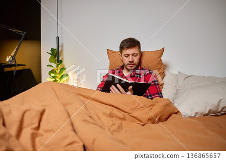 Man sitting in bed and reading book under blanket in bedroom Man sitting in bed and reading book under blanket in bedroom 136865657