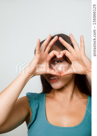 Woman making a heart shape with her hands while smiling at the camera in a simple setting during the daytime Woman making a heart shape with her hands while smiling at the camera in a simple setting during the daytime 136866129