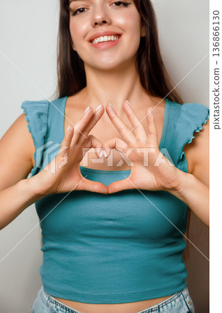 Woman making a heart shape with her hands, smiling at the camera in a bright indoor setting during daytime 136866130