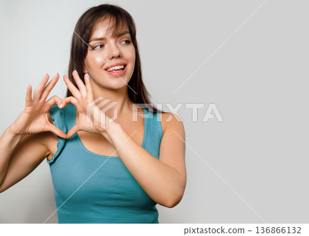 Woman makes heart shape with her hands in a studio setting while smiling in a simple blue shirt during daytime Woman makes heart shape with her hands in a studio setting while smiling in a simple blue shirt during daytime 136866132