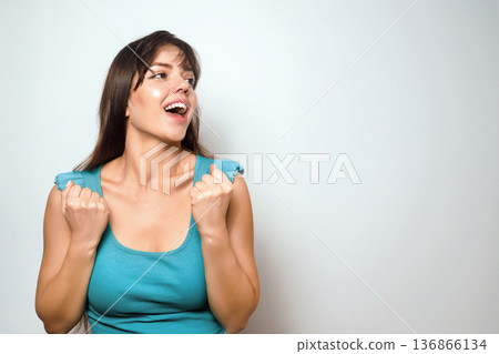 Woman shows excitement and joy while standing against a plain background during a casual indoor setting Woman shows excitement and joy while standing against a plain background during a casual indoor setting 136866134