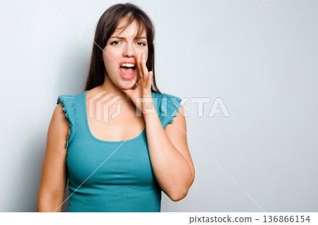 Woman shouting with hand near mouth while standing against plain background in indoor setting during daytime 136866154