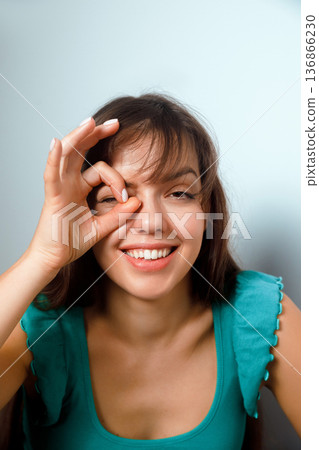 Woman makes a silly gesture with her hand over one eye while smiling and sitting in a casual setting against a light background Woman makes a silly gesture with her hand over one eye while smiling and sitting in a casual setting against a light background 136866230