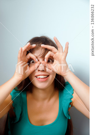 Young woman makes funny gesture with hands while smiling indoors in bright light against a plain wall in a casual setting Young woman makes funny gesture with hands while smiling indoors in bright light against a plain wall in a casual setting 136866232