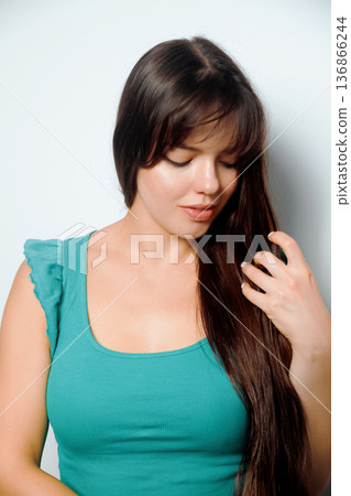 Young woman with long hair plays with her hair while looking down in a simple indoor setting during daytime 136866244