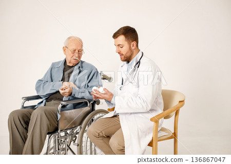 Senior gentleman on a wheelchair isolated on a white background. Man wearing blue t-shirt and doctor wearing white medical gown. Doctor talking with man and give him medicines. Senior gentleman on a wheelchair isolated on a white background. Man wearing blue t-shirt and doctor wearing white medical gown. Doctor talking with man and give him medicines. 136867047