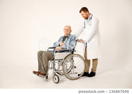 Senior gentleman on a wheelchair isolated on a white background. Man wearing blue t-shirt and doctor wearing white medical gown. Doctor talking with man. Senior gentleman on a wheelchair isolated on a white background. Man wearing blue t-shirt and doctor wearing white medical gown. Doctor talking with man. 136867048