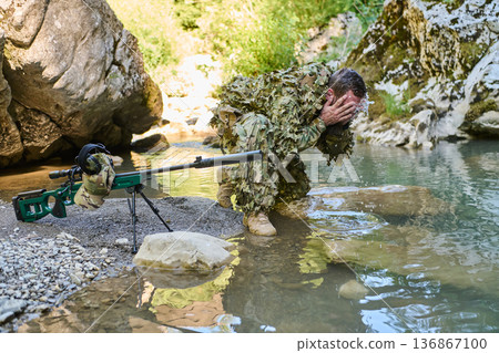 Soldier In Ghillie Suit Cleansing Face At Riverbank Next To Sniper Rifle During Field Exercise 136867100