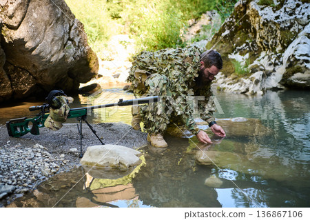 Camouflaged Sniper Soldier Kneeling By River With Precision Rifle Collecting Water In Wilderness Camouflaged Sniper Soldier Kneeling By River With Precision Rifle Collecting Water In Wilderness 136867106