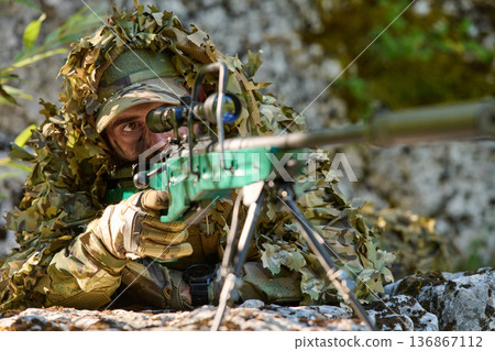 Soldier Sniper in Ghillie Camouflage Aiming a Precision Rifle From Prone Position in Woodland 136867112